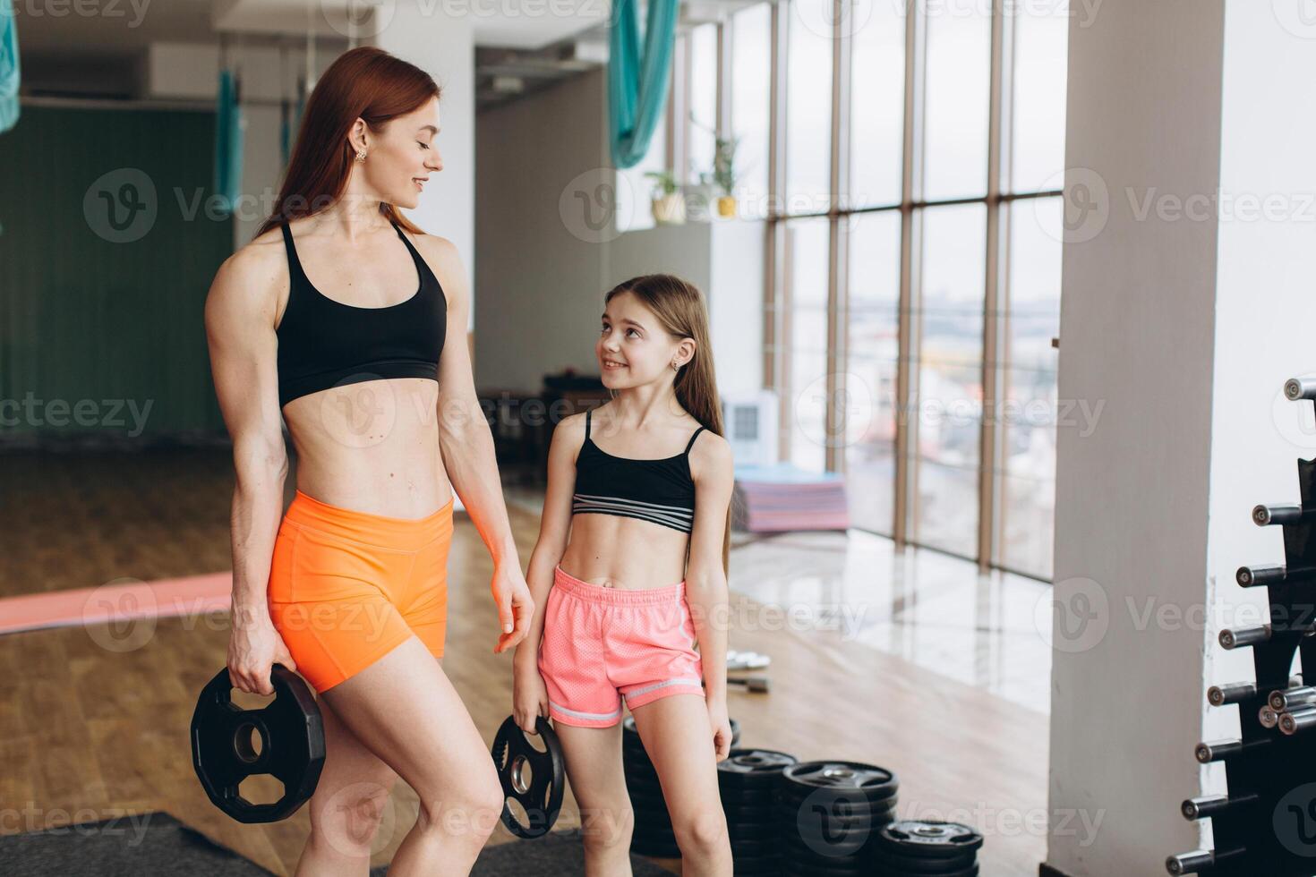 Mom and Daughter are doing exercises. Family in a gym. Little girl with strong mother are training with pancakes rods photo