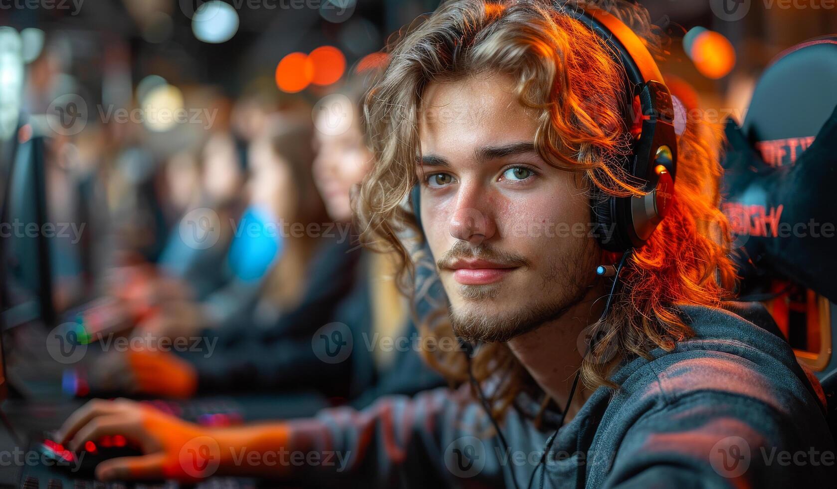 A man with long hair is wearing headphones and looking at the camera. He is in a room with other people, some of whom are also wearing headphones. Scene is casual and relaxed photo