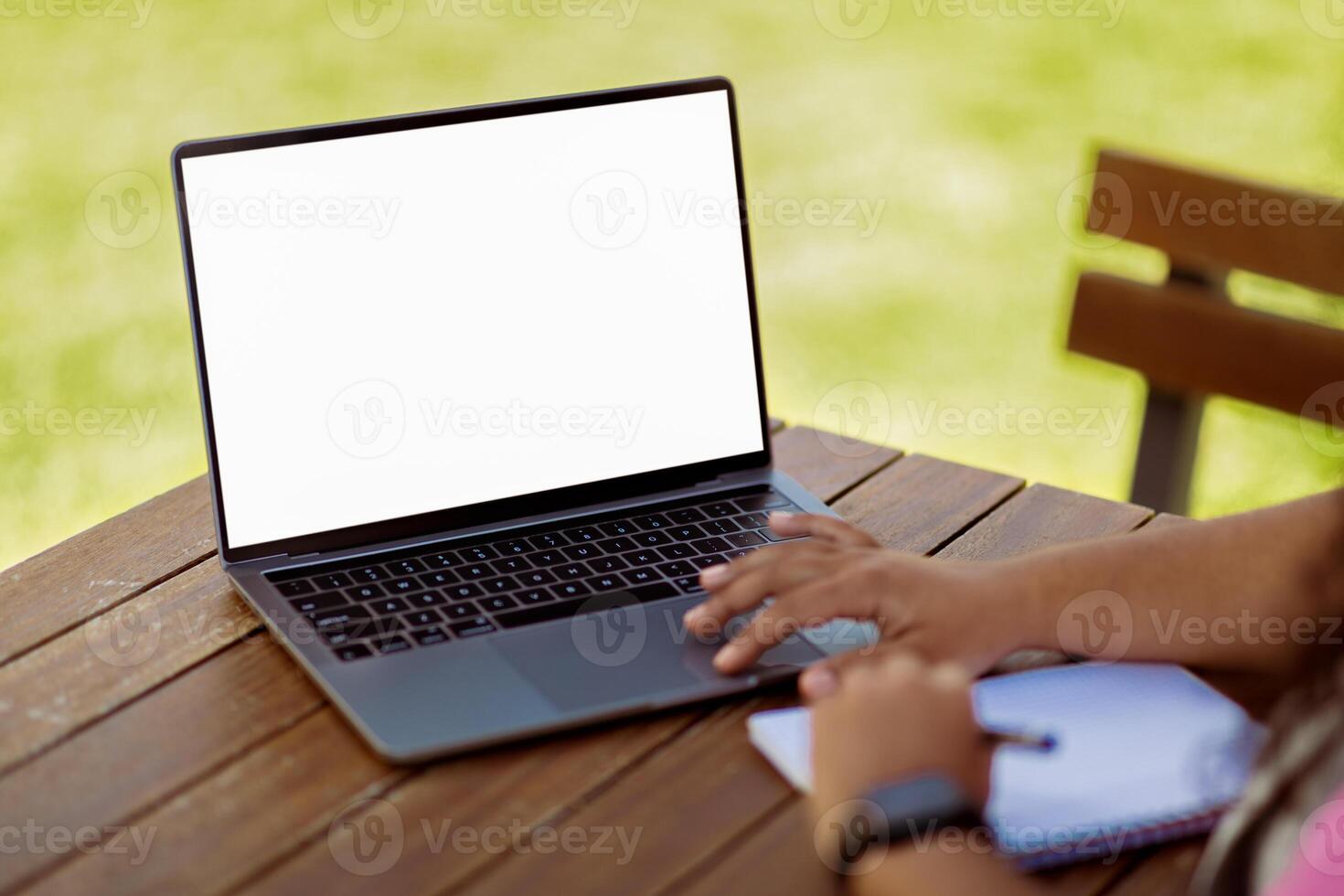 A person is seated at a wooden table, typing on a laptop under the sun, with a notepad beside them. The vibrant green grass in the background enhances the relaxing atmosphere of the workspace. photo