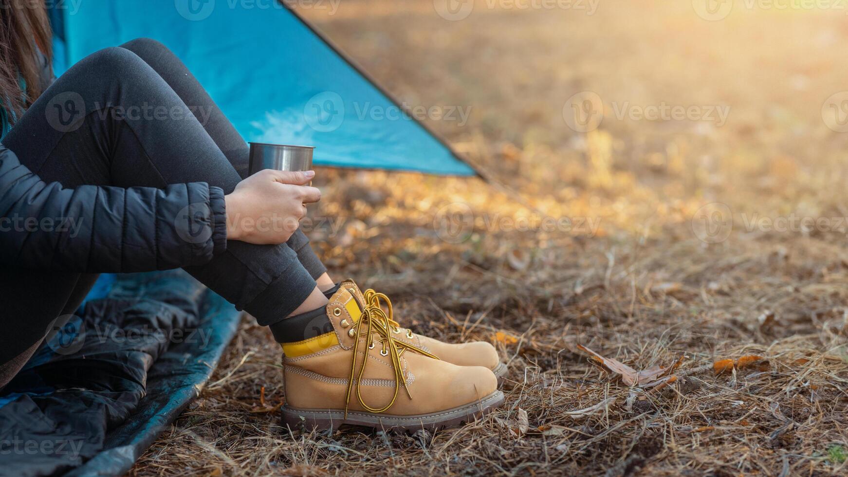 Relaxation concept. Legs in boots of woman camping in forest, holding cup, empty space photo