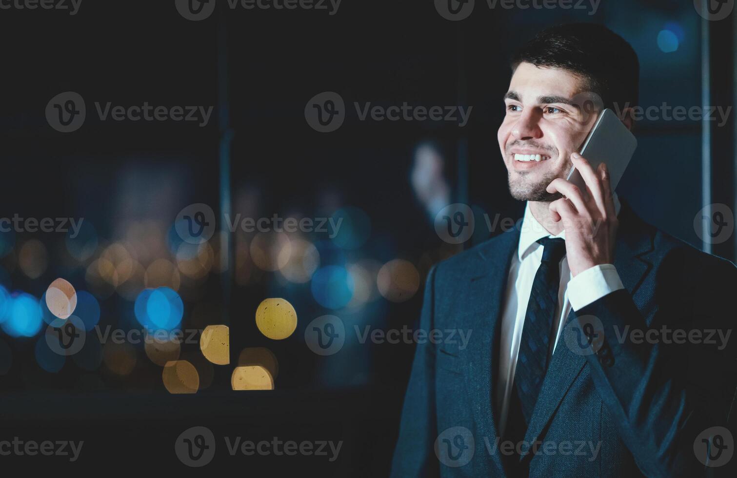 Smiling Entrepreneur Talking On Phone Standing Near Window With City View In Modern Office At Night. Business Communication. Copy Space photo