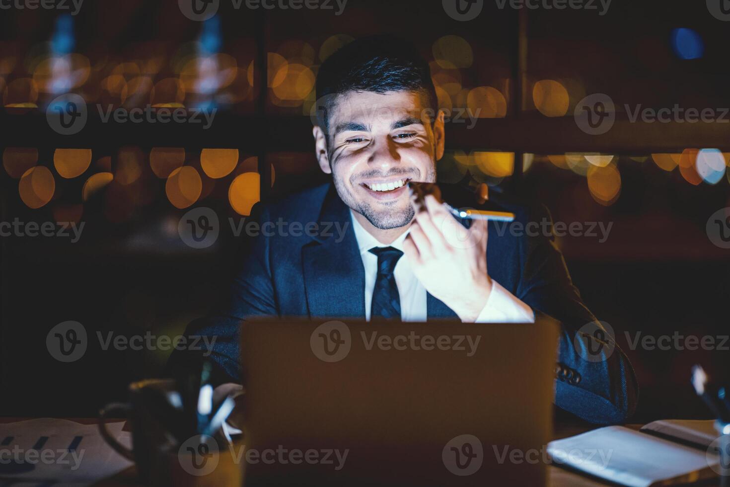 Office Manager Guy Using Voice Assistant On Smartphone Working On Laptop In Modern Office Late At Night. Selective Focus photo
