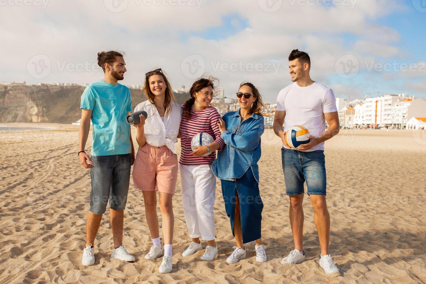 A group of five friends stand on a sandy beach enjoying each other company. They are dressed casually and are engaged in lively conversation while holding volleyballs. photo