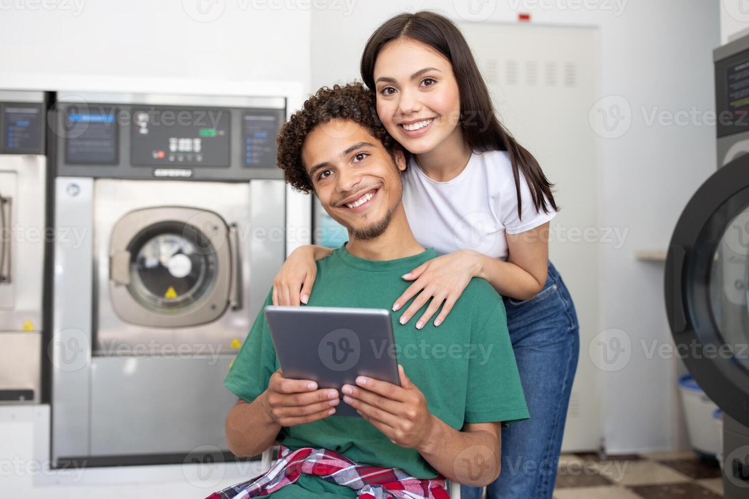A young couple relaxes together in a laundromat, with the man seated and holding a tablet. The woman stands behind him, smiling and enjoying their time while waiting for laundry. photo