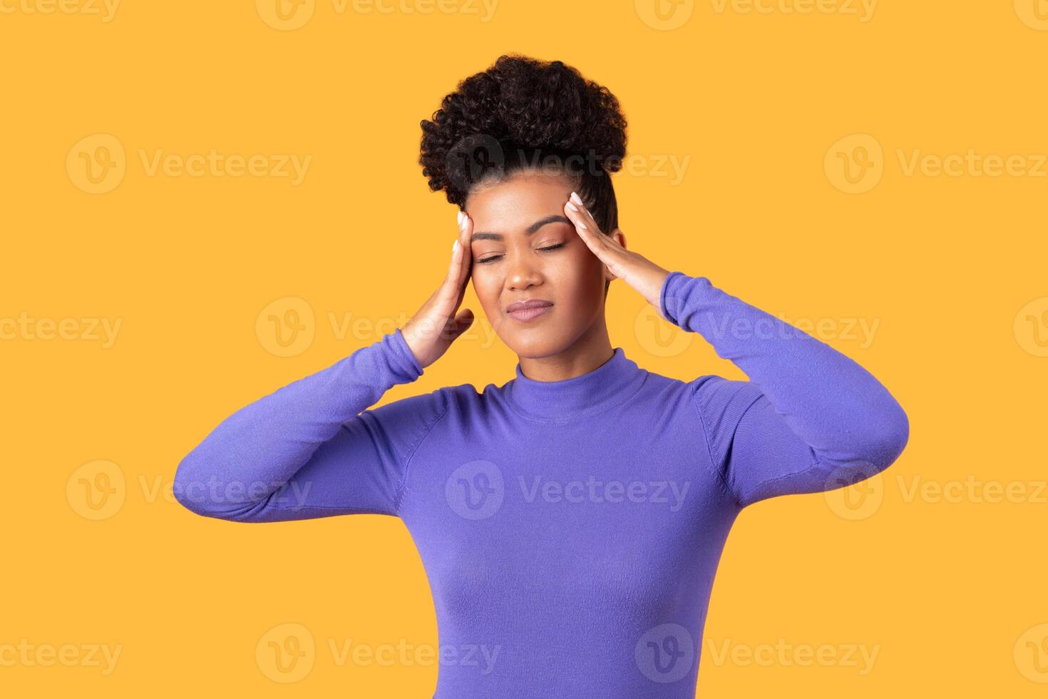 A young woman in a purple long-sleeve top practices stress relief by placing her hands on her temples, appearing focused on calming her mind in a vibrant yellow setting. photo