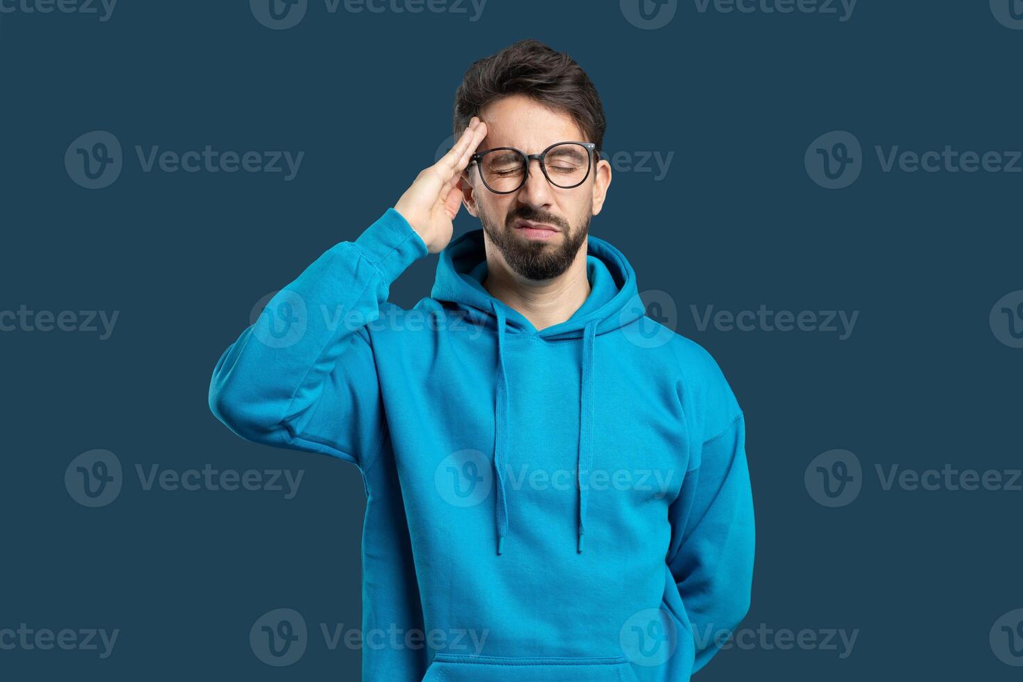 A young man wearing a blue hoodie and glasses expresses frustration while standing against a solid blue background. His hand rests on his forehead as he contemplates. photo