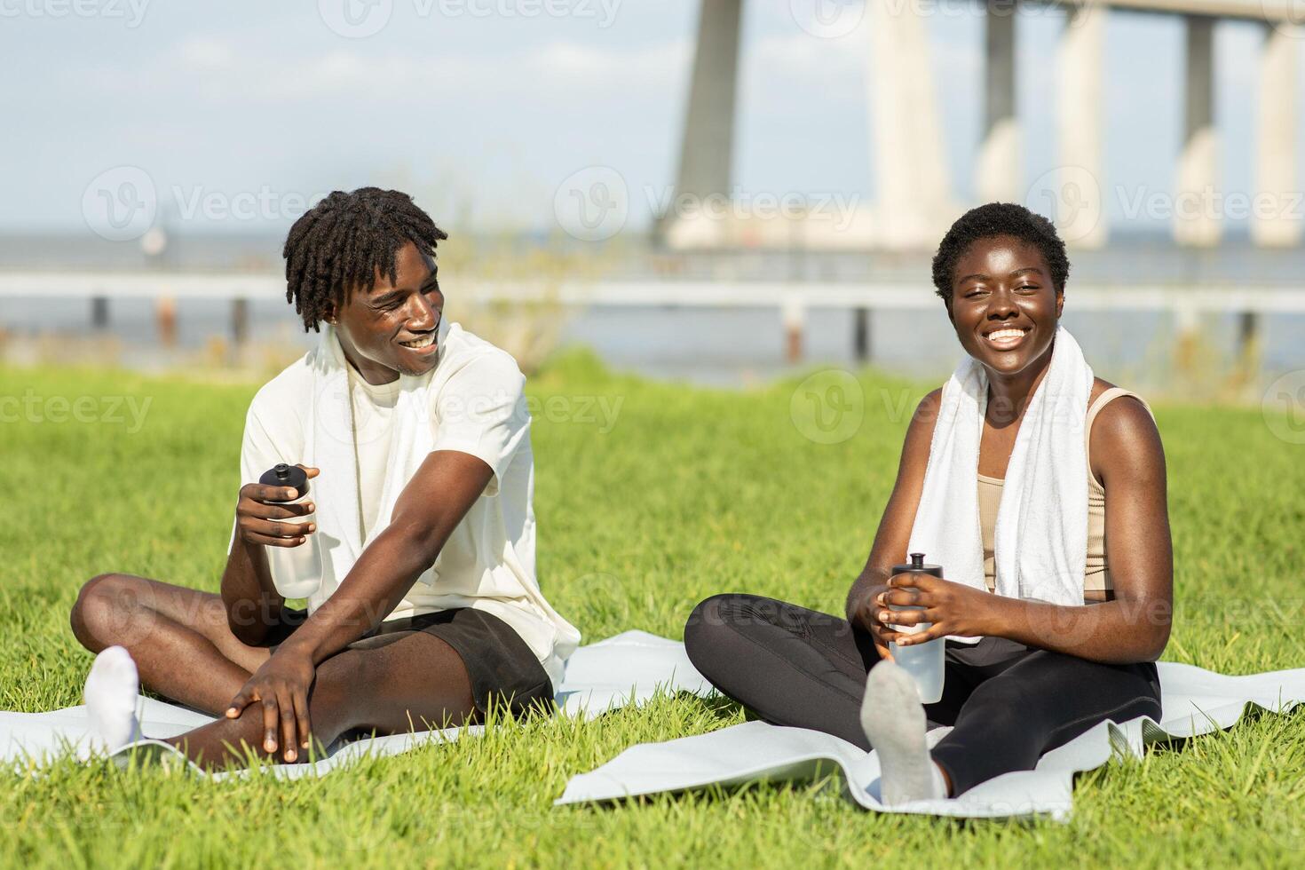 Two African American friends relax on yoga mats by the water, enjoying a sunny day. They smile and sip from water bottles after a yoga session, surrounded by grass and calm scenery. photo