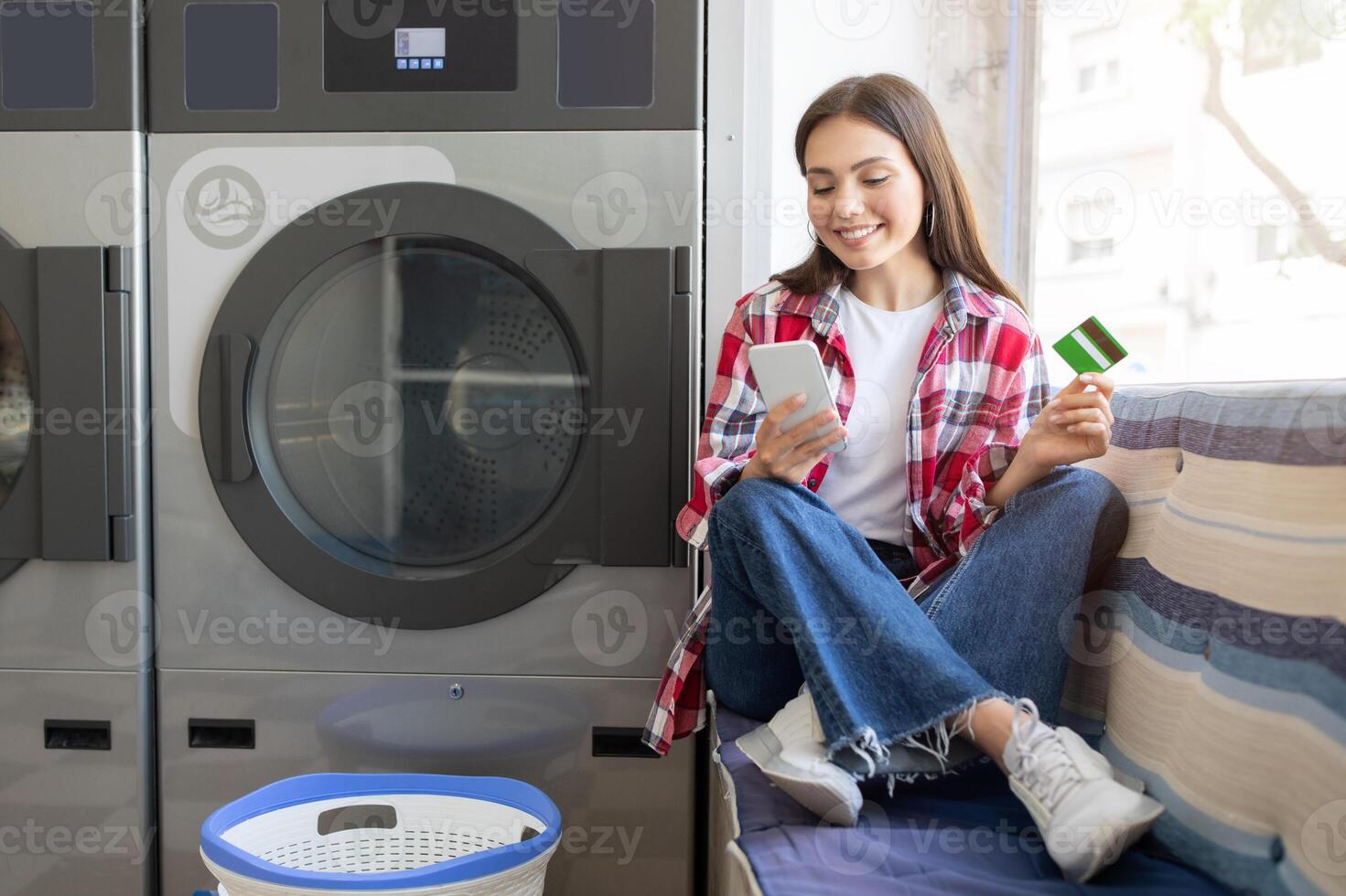 A young woman sits comfortably on a bench in a laundromat, using her smartphone while holding a payment card. She smiles as she waits for her laundry to finish. photo