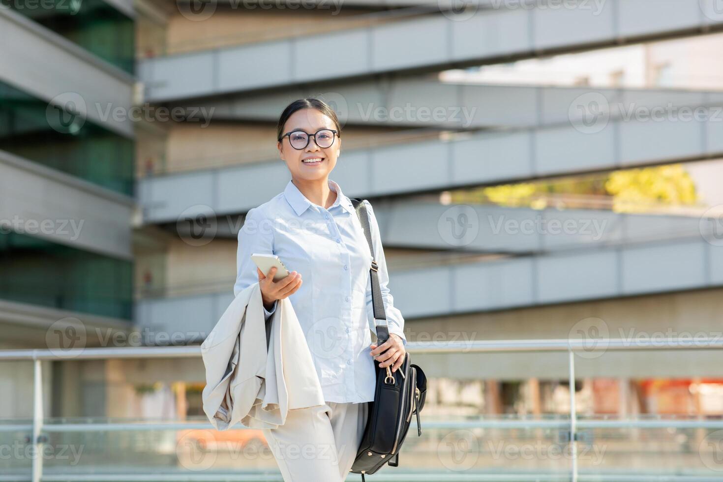 Asian woman dressed in smart attire stands confidently in an urban environment, holding a smartphone and smiling. The backdrop features contemporary architectural elements that enhance the setting. photo