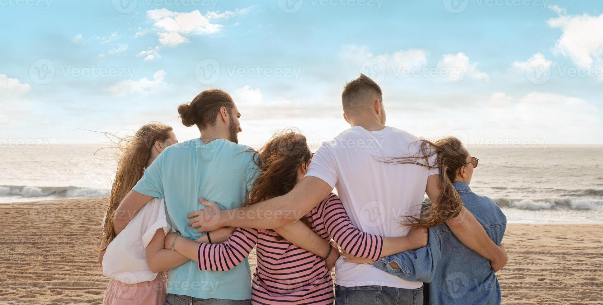 A group of friends stands together on the beach, overlooking the ocean as the sun sets, with their arms around each other, capturing a moment of camaraderie and joy, back view photo