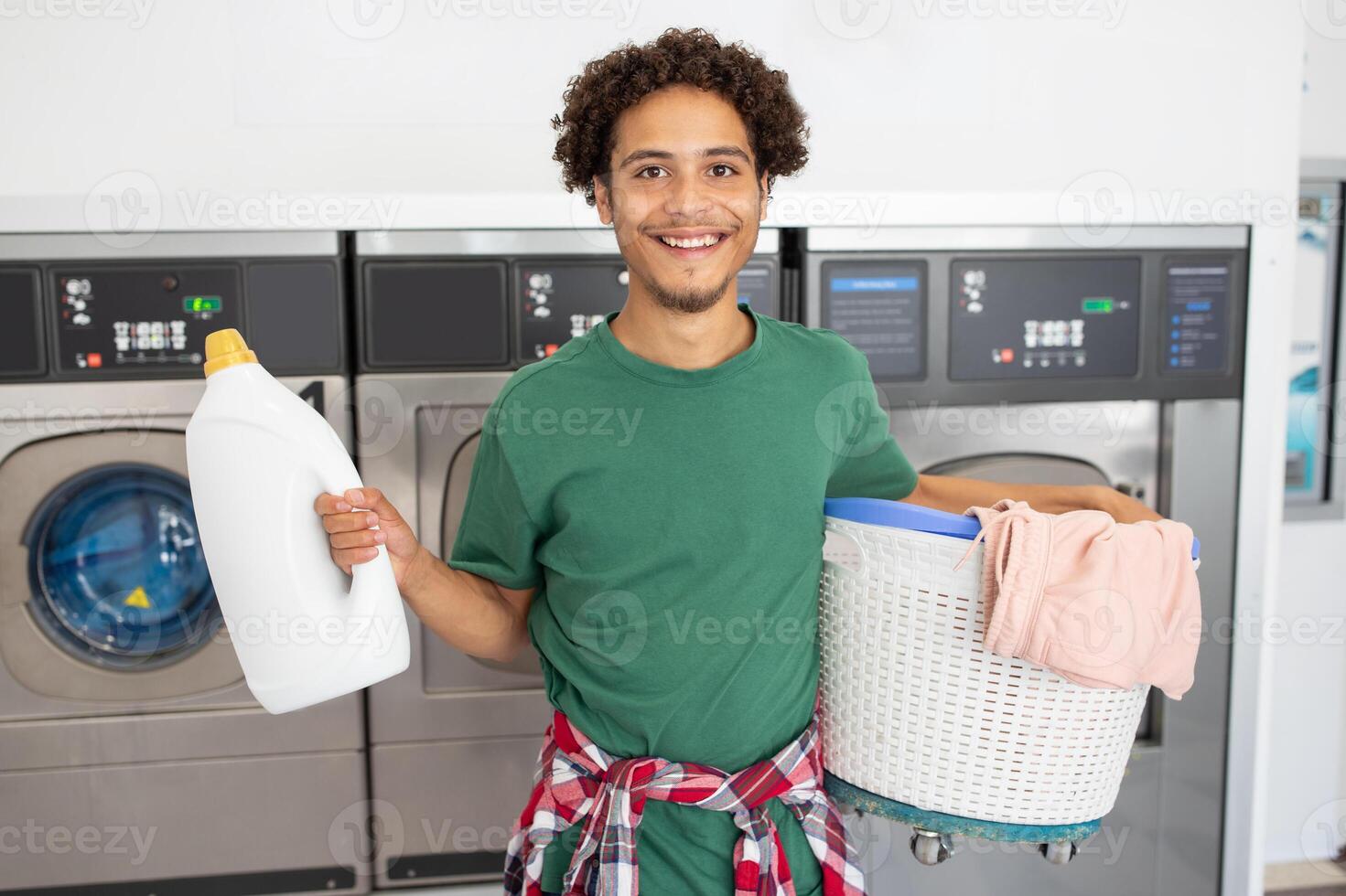 A young man stands in a laundromat, holding a laundry basket filled with clothes in one hand and a bottle of detergent in the other. He has a cheerful expression while ready to wash. photo