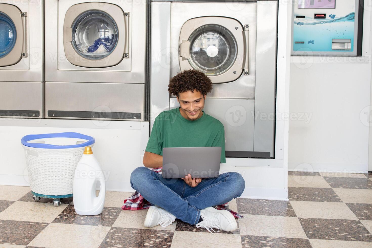 A young man sits on the floor of a laundromat, using a laptop for studies while waiting for his clothes to wash. Nearby, there is a laundry basket and detergent bottle. photo