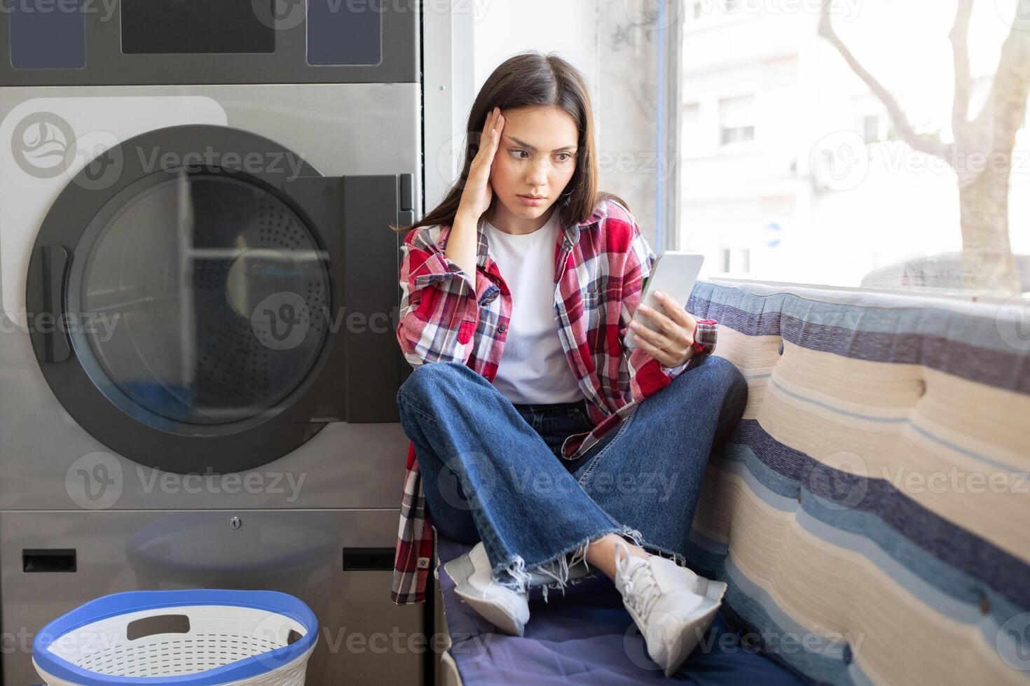 A young woman sits on a bench in a laundromat, looking stressed while checking her phone. She has a plaid shirt on and appears to be waiting for her laundry to finish. photo