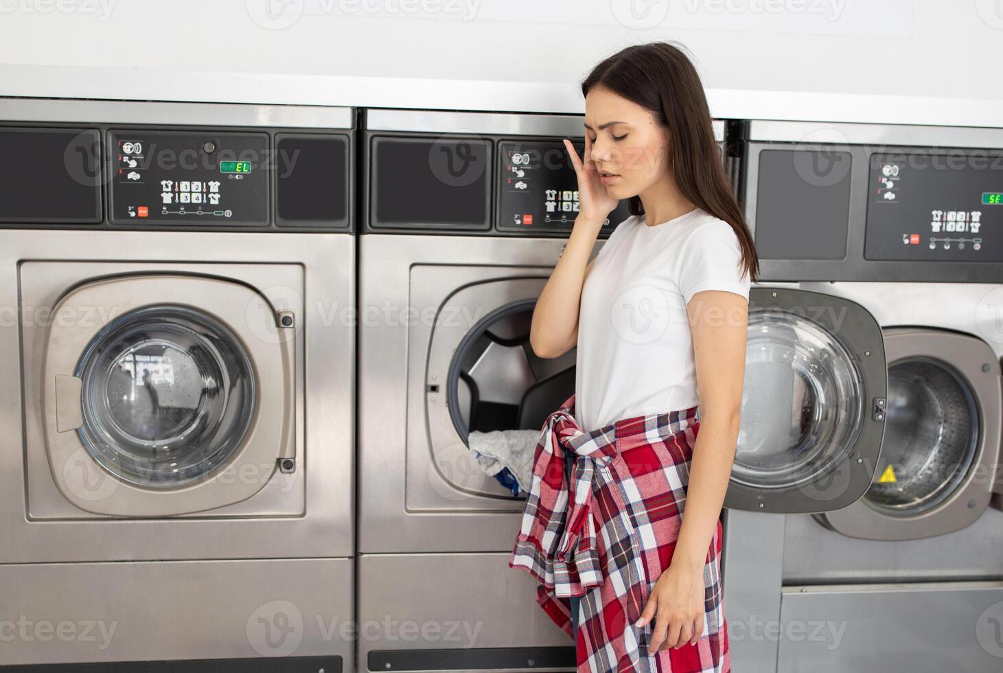 A young woman stands beside laundry machines, visibly frustrated as she waits for her clothes to finish washing in a busy laundromat during the weekend, have headache photo
