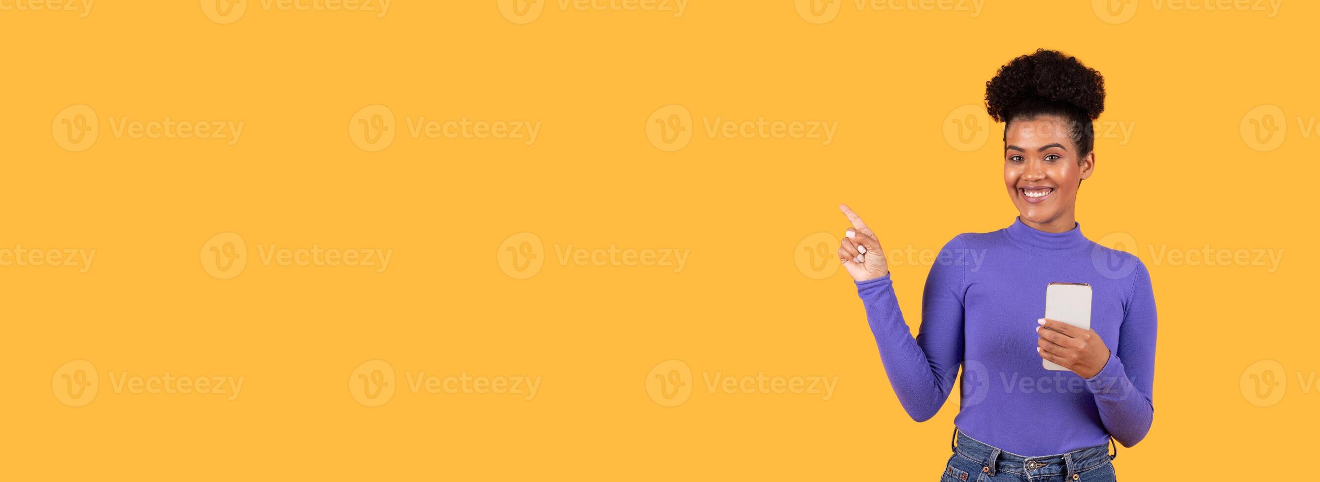 A joyful woman in a purple top stands against a vibrant yellow backdrop, holding a smartphone in one hand while playfully pointing at copy space, creating a cheerful and engaging atmosphere. photo
