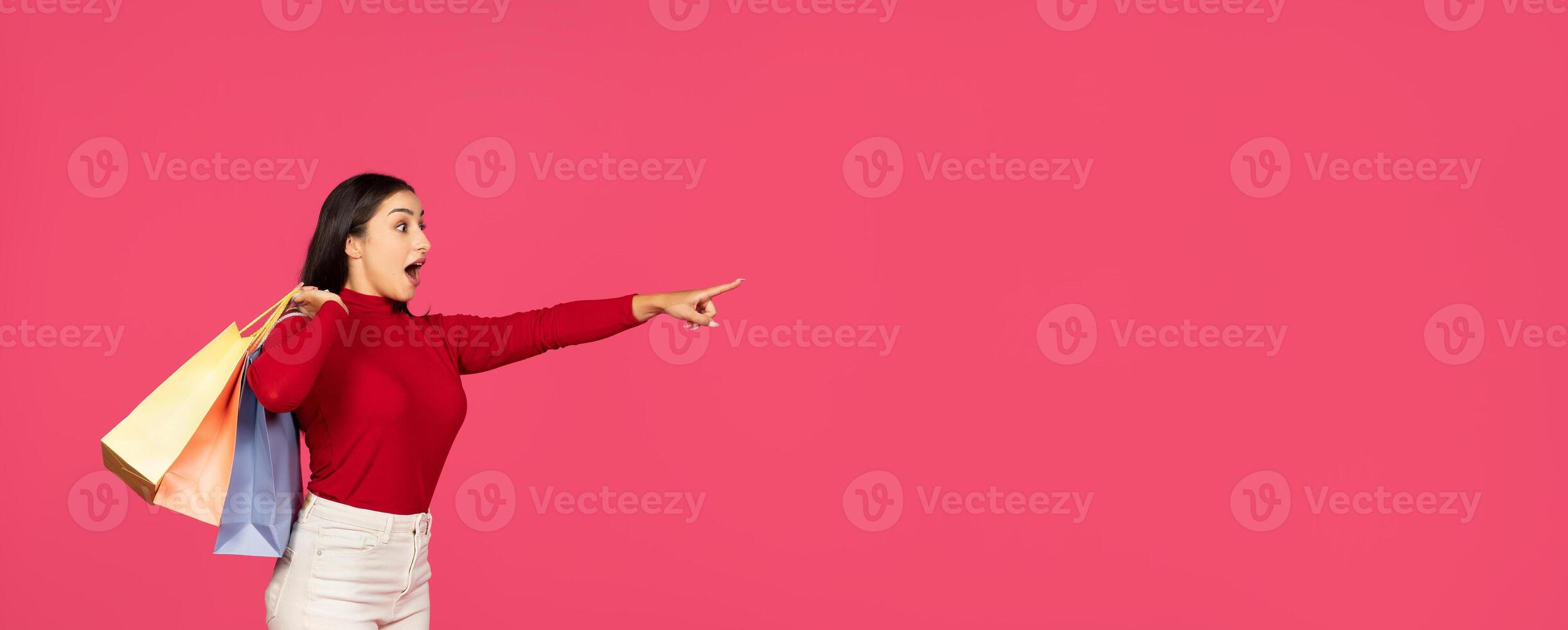 A woman wearing a red sweater displays an enthusiastic expression as she points towards copy space, holding colorful shopping bags in her other hand, set against a vibrant pink backdrop. photo