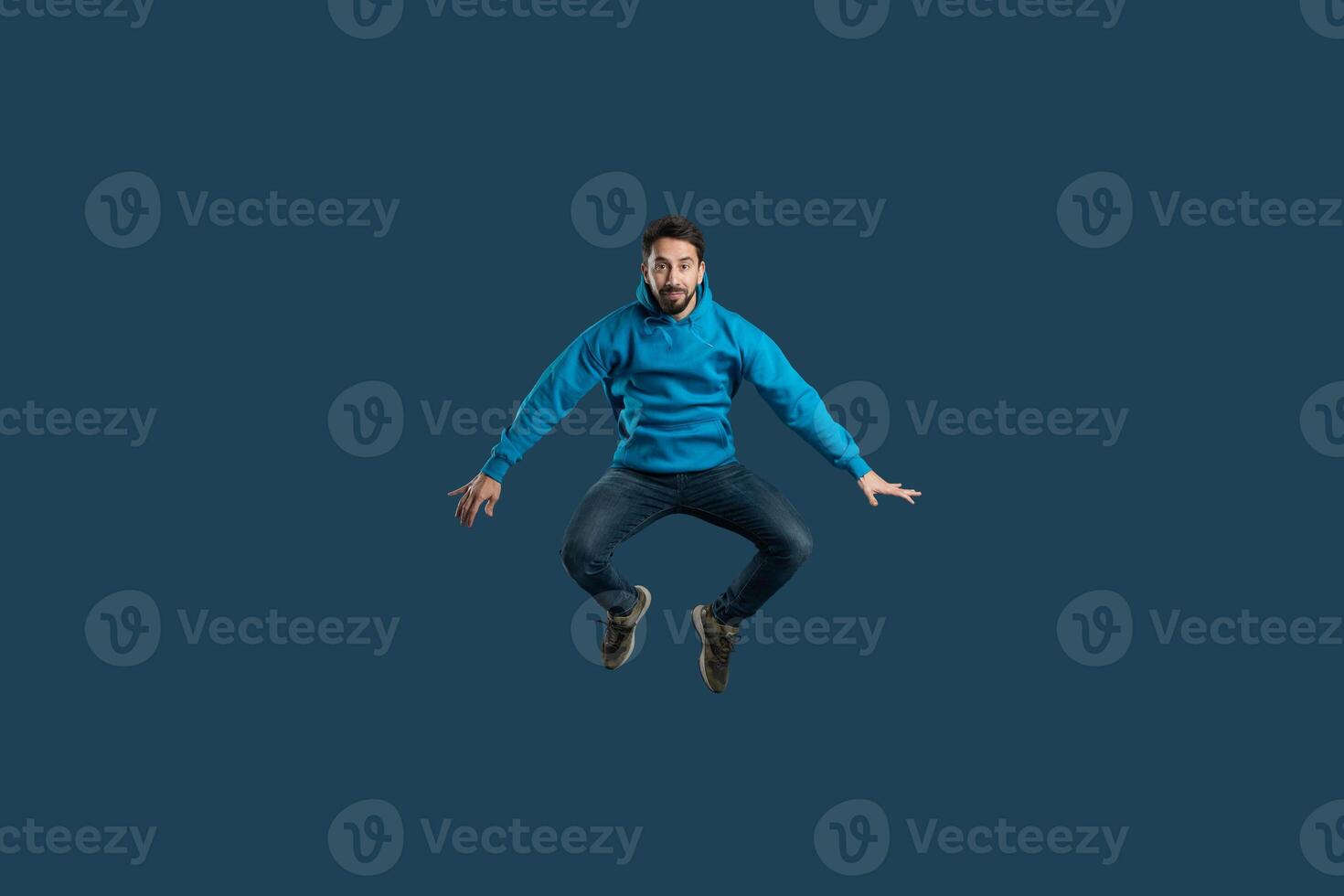 A man in a blue hoodie is mid-air, leaping with joy. The dark background emphasizes his vibrant outfit and energetic pose, showcasing an expression of excitement and freedom. photo