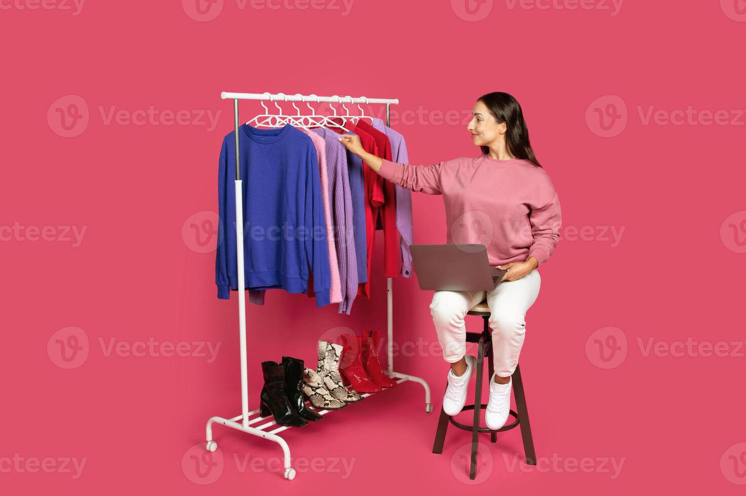 A woman sits on a stool, working on a laptop while examining clothing on a rack. The vibrant background enhances the bright colors of the apparel displayed, setting a lively atmosphere. photo