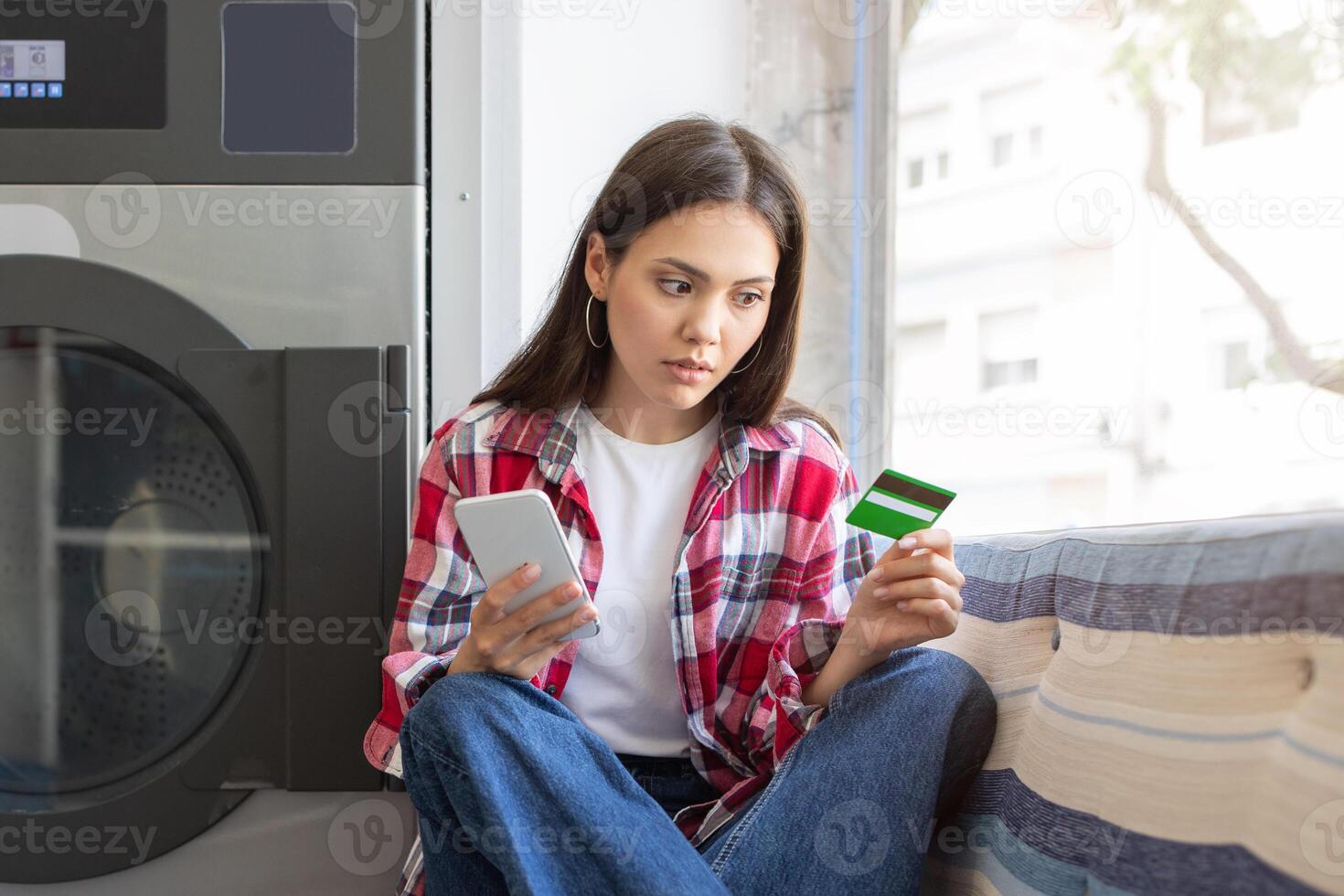 A young woman sits in a brightly lit laundromat, holding a smartphone in one hand and a credit card in the other, looking thoughtful as she prepares to make a payment. photo