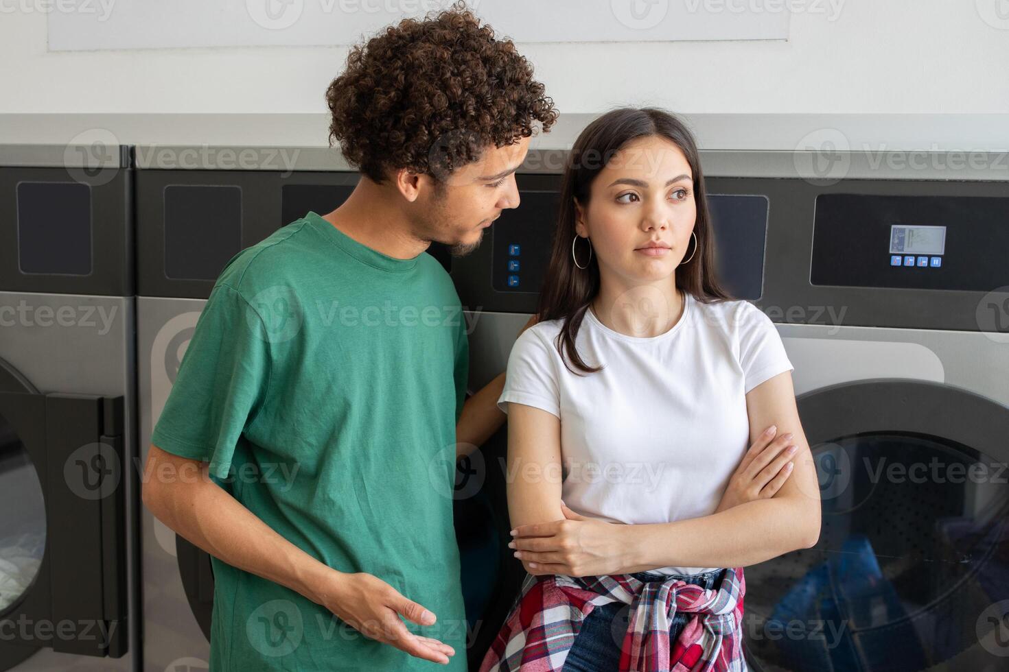 A young man and woman stand in a laundromat. The man appears to be discussing something serious with the woman, who looks thoughtful and slightly distressed while crossing her arms. photo