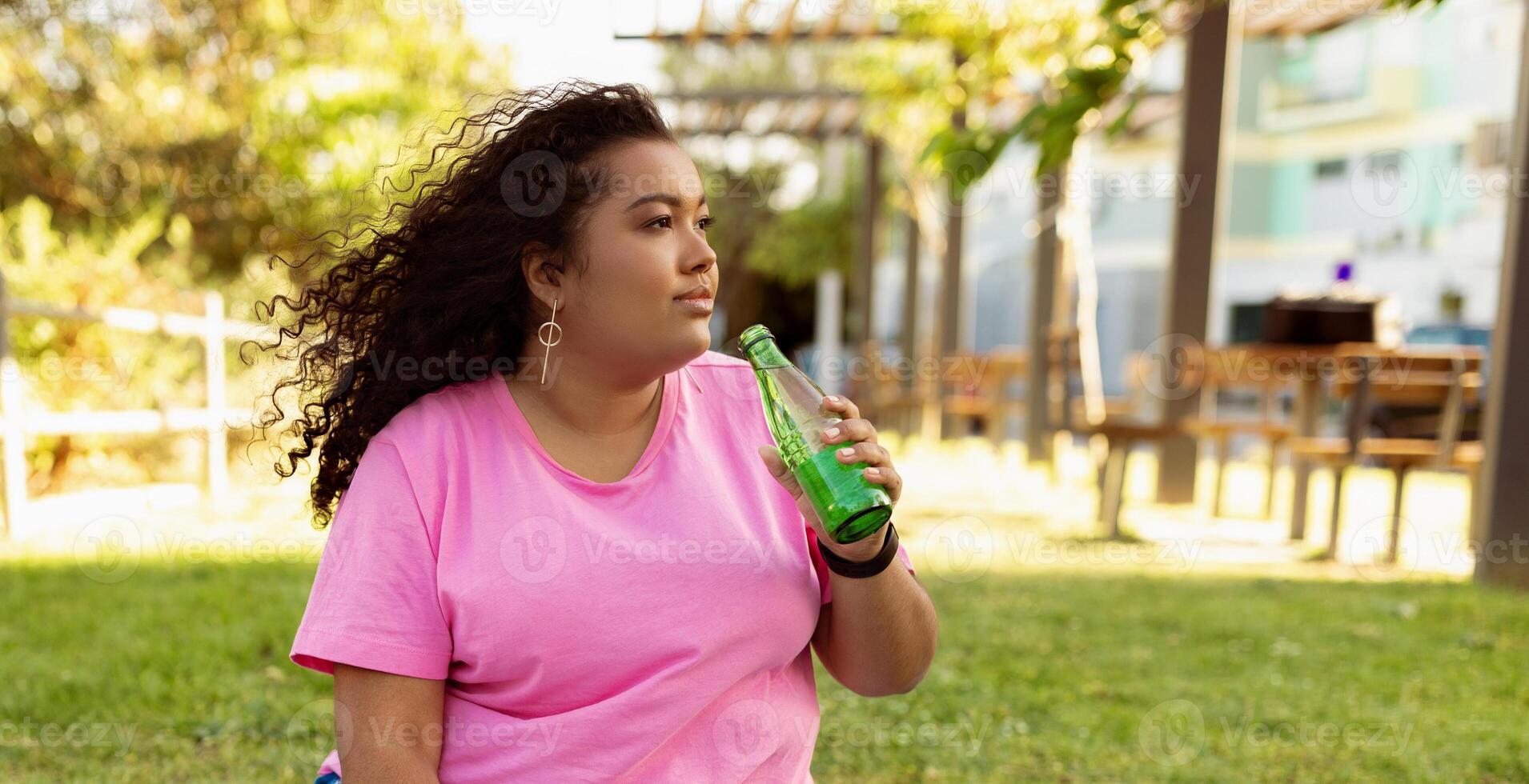 A woman sits on the grass in a park, basking in the sunlight while sipping from a green beverage. Surrounding her are trees and benches, creating a relaxing environment. photo