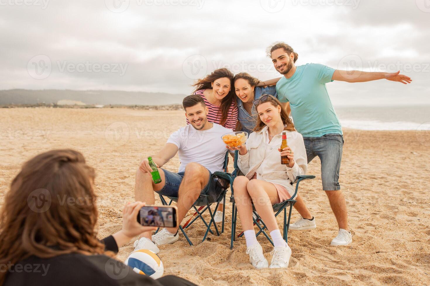A group of five friends gathers on the beach, sitting in chairs and posing cheerfully for a photo. They hold drinks and snacks, creating a relaxed and joyful atmosphere by the shoreline. photo