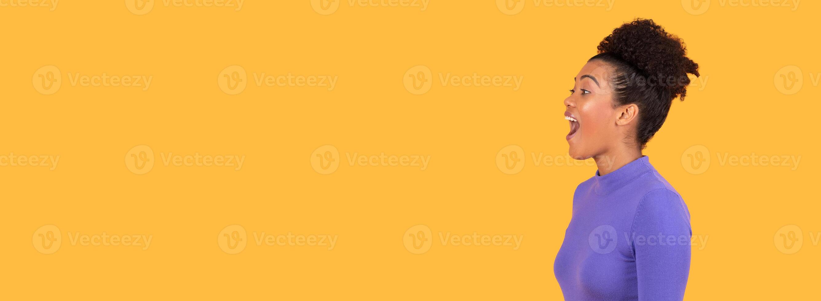 A woman with a curly hairstyle exclaims in delight, her face lit up with joy. She stands indoors against a vibrant yellow backdrop, emphasizing her lively expression, panorama with copy space photo