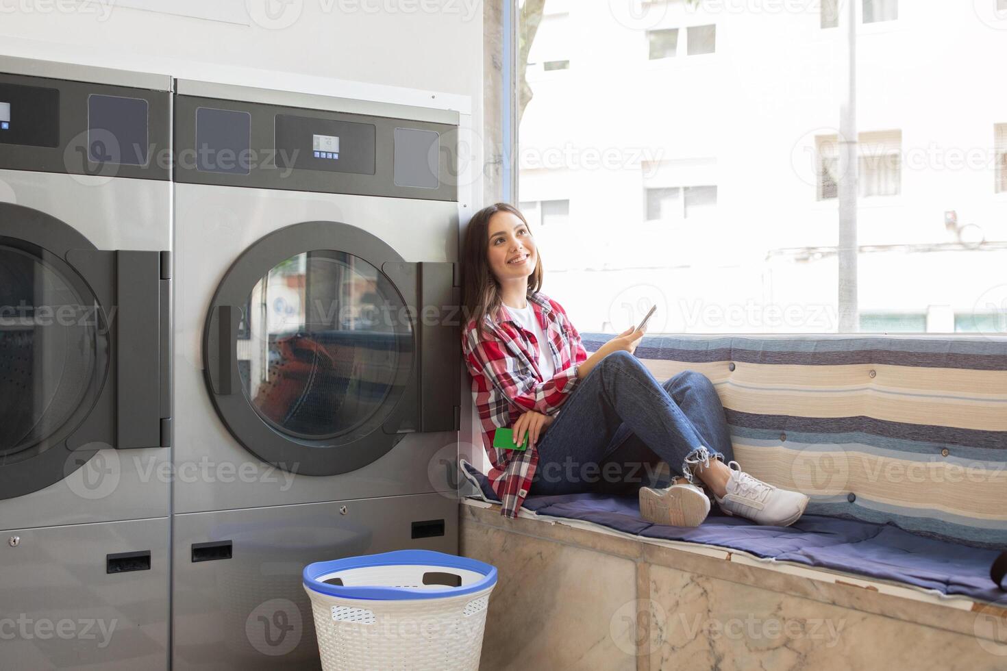 A young woman sits comfortably by a window in a laundromat, using her phone as she enjoys her time while waiting for her laundry. Bright light streams in from outside. photo