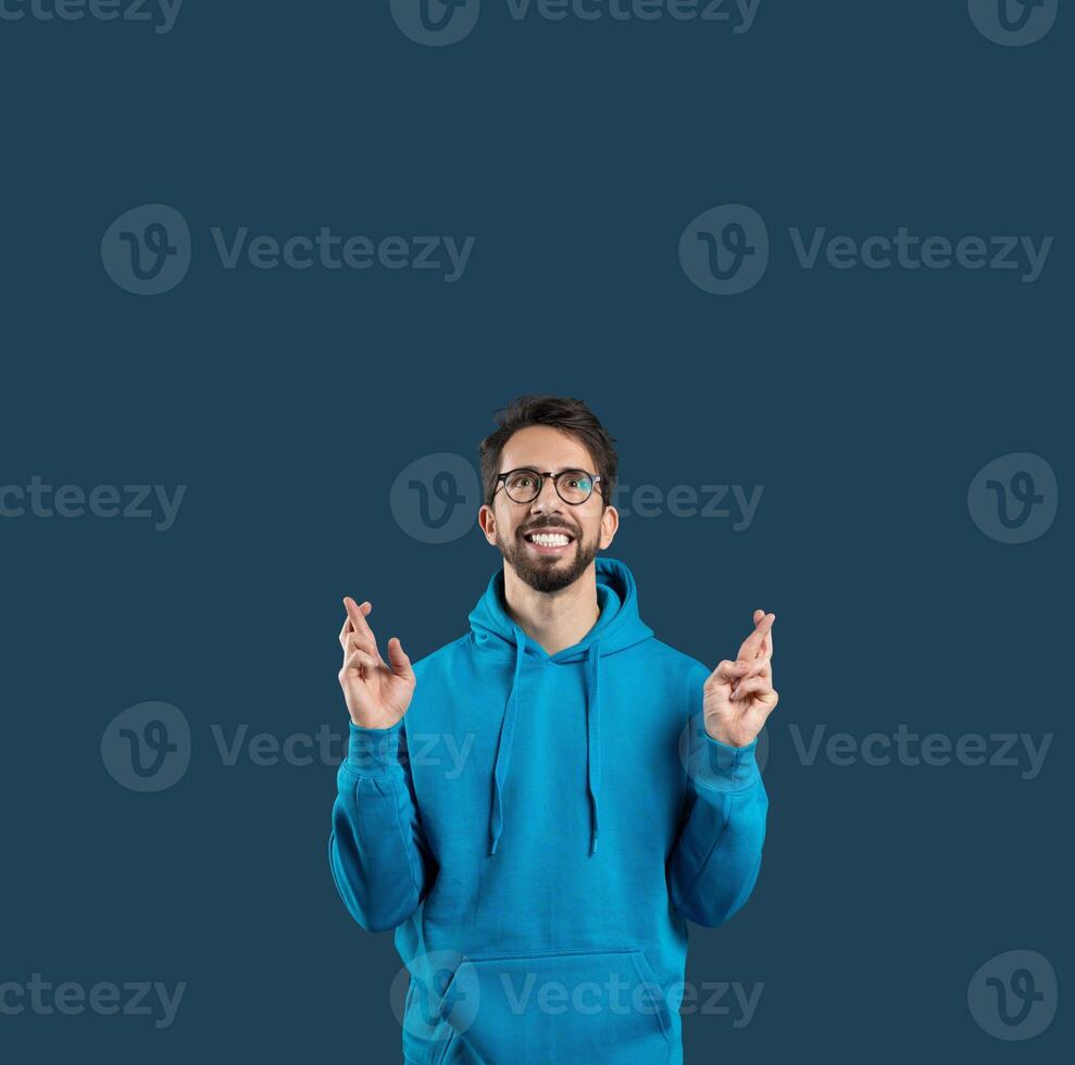 A young man with glasses is wearing a bright blue hoodie, smiling and crossing his fingers as a gesture of hope or good luck. The background is a solid dark color, copy space photo