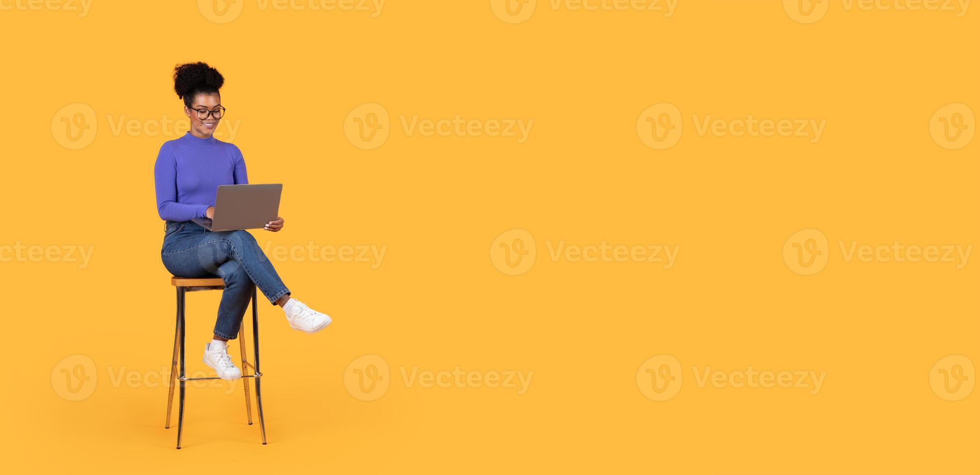 A woman with curly hair, wearing glasses, smiles while seated on a stool, working on her laptop. Bright yellow background creates a cheerful atmosphere, enhancing her focused expression, copy space photo