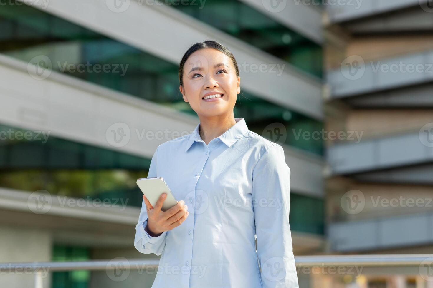 Asian woman stands outdoors in an urban setting, smiling and holding a smartphone. She enjoys a pleasant moment, surrounded by modern buildings and a bright sky. photo
