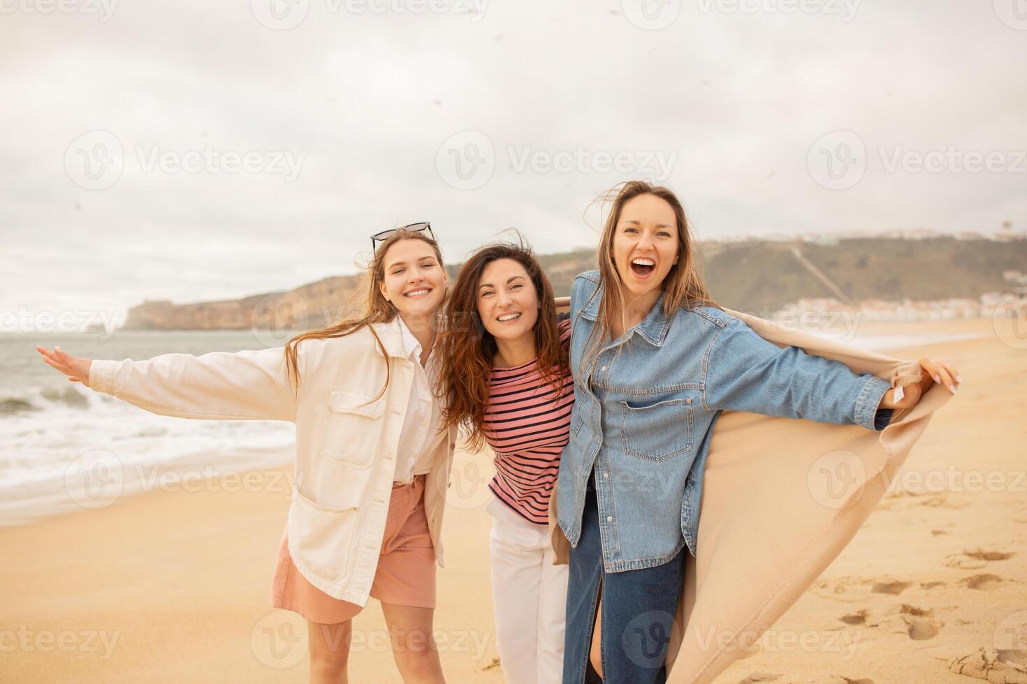 Three friends celebrate a joyful day at the beach, laughing and embracing each other as waves crash gently in the background. The sandy shore creates a perfect summer atmosphere. photo