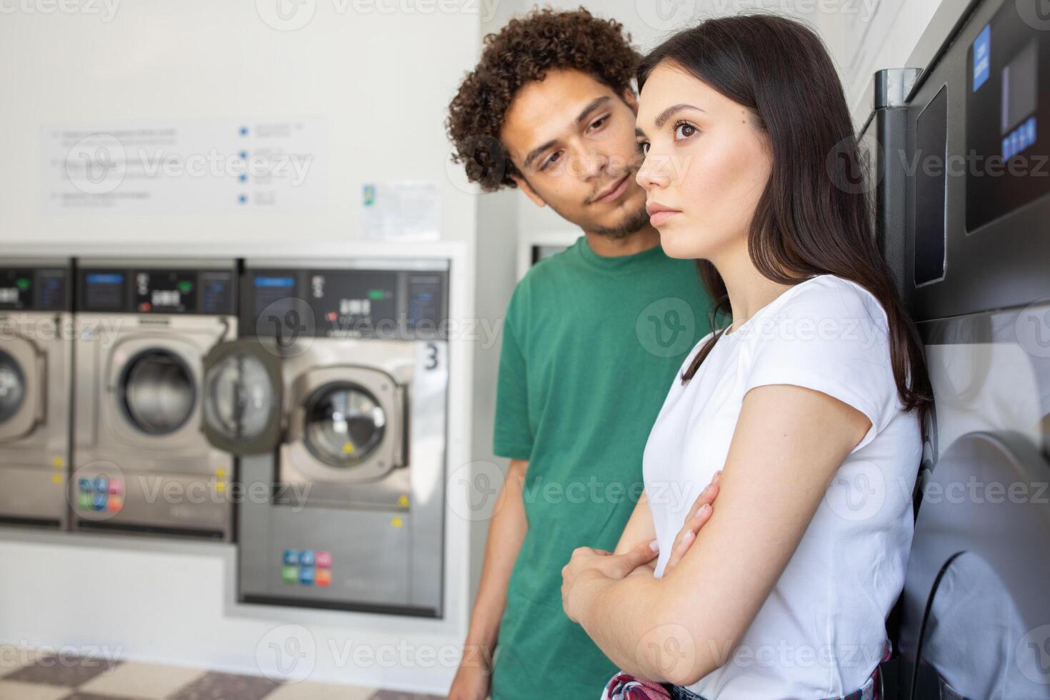 A young couple stands in a laundromat, one person looks at the other with interest while the other appears contemplative, reflecting the mood of their conversation during laundry time. photo