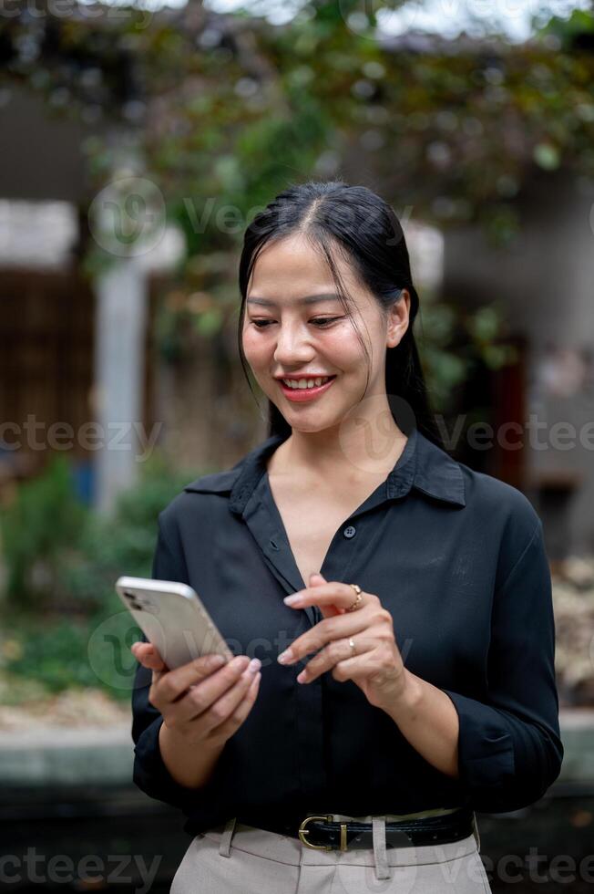 A charming Asian businesswoman is reading text messages on her smartphone in a garden space. photo