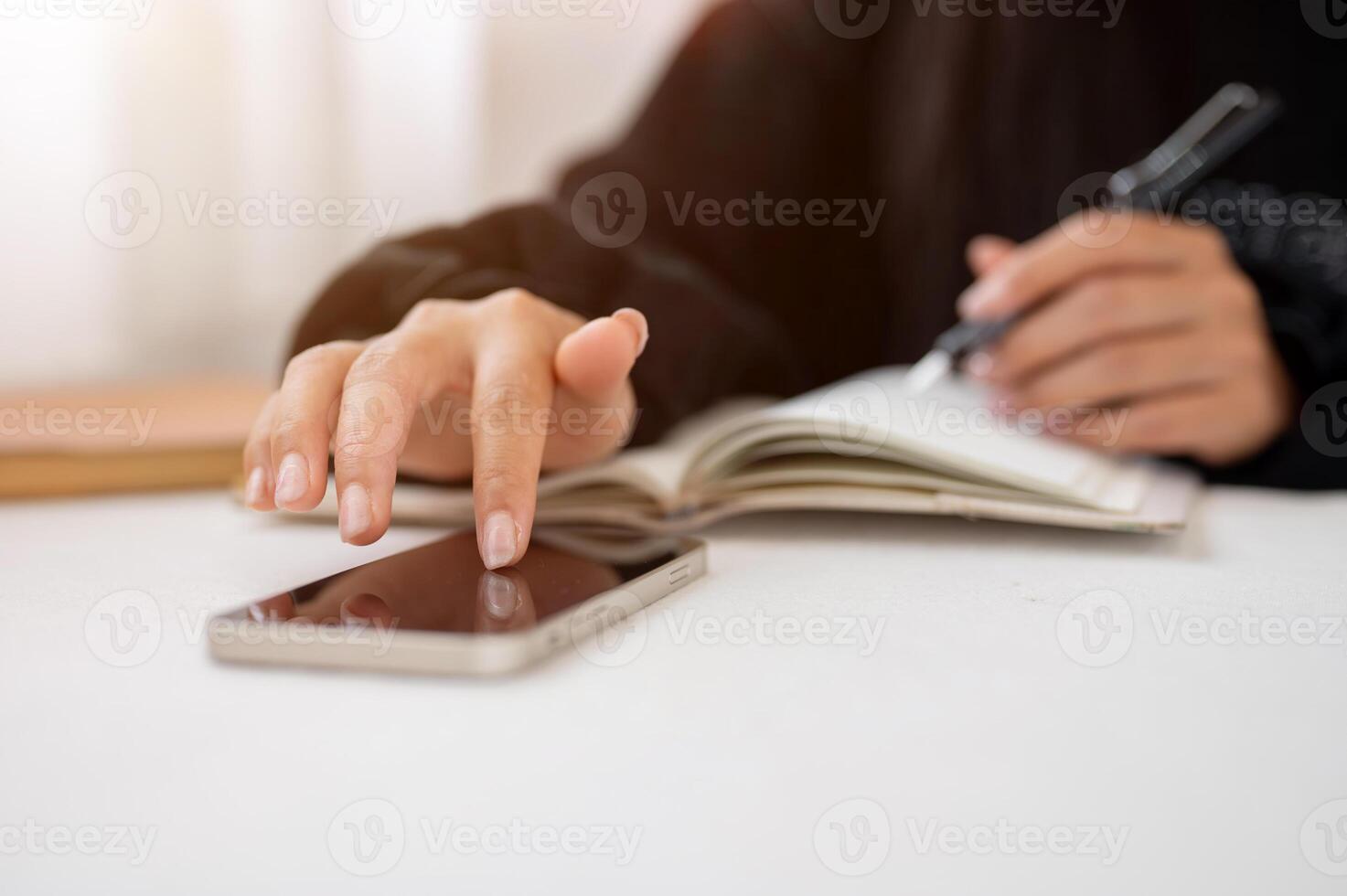 A close-up of a woman writing in a book and using her smartphone while sitting at a table indoors. photo