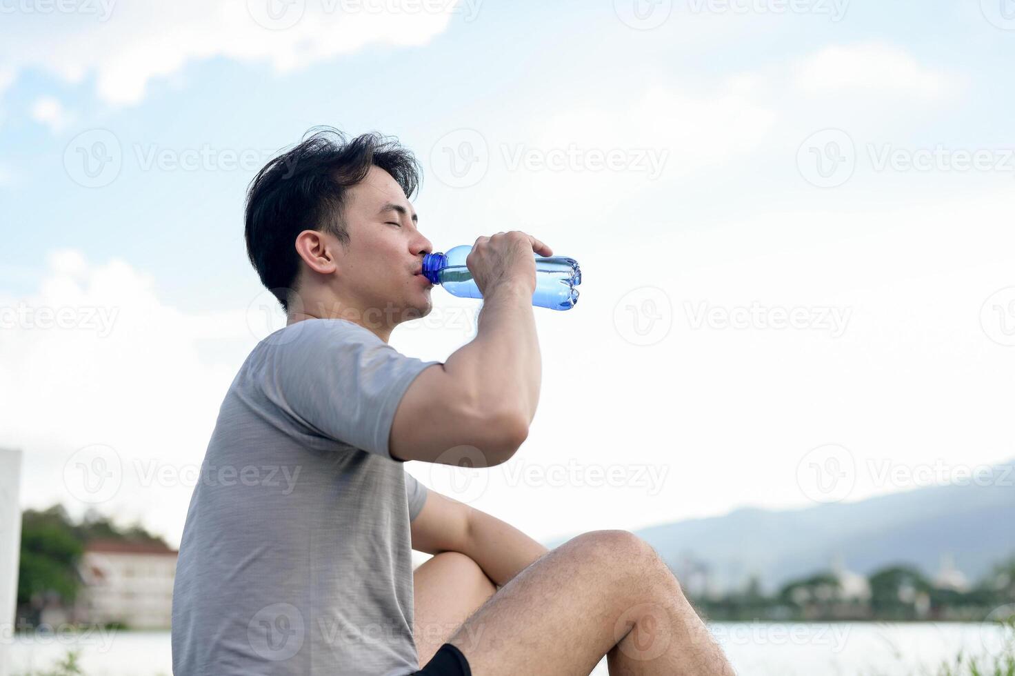 A tired, sporty Asian man in sportswear sits by the lake, drinking to refresh himself after a run. photo