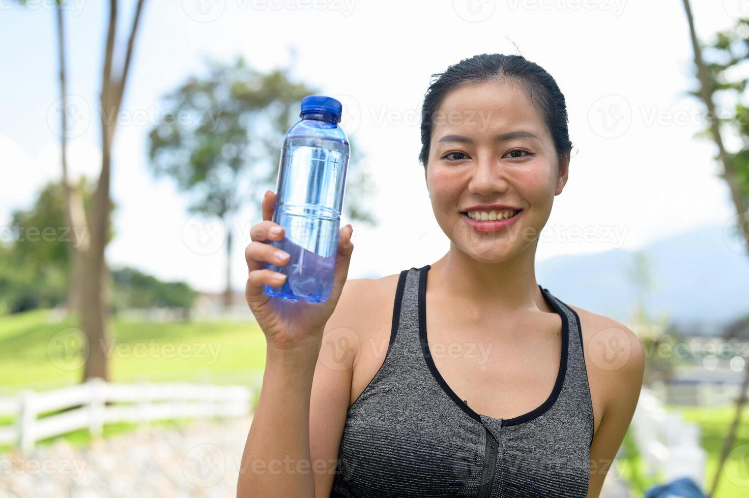 A fit Asian woman in sportswear is holding up her water bottle, standing in a green park. photo