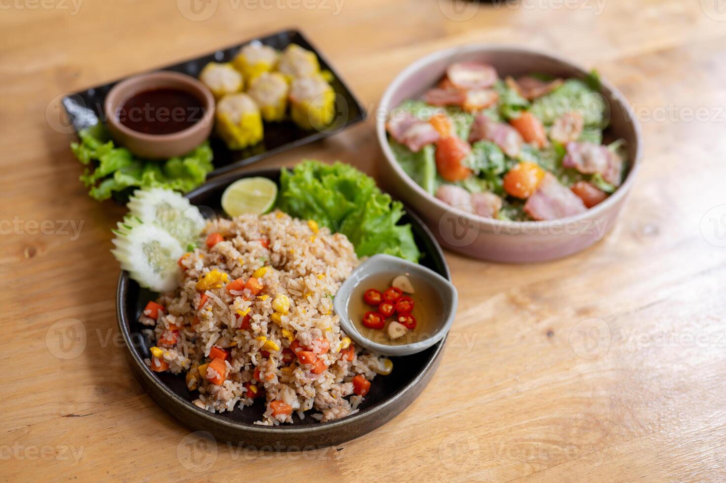 A wooden dining table featuring a plate of fried rice, a salad bowl, and a plate of steamed dumpling photo