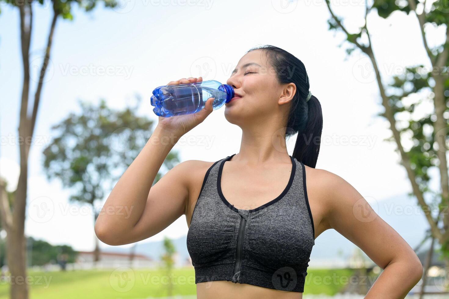 A fit woman in sportswear drinks from a water bottle, quenching her thirst after jogging in a park. photo