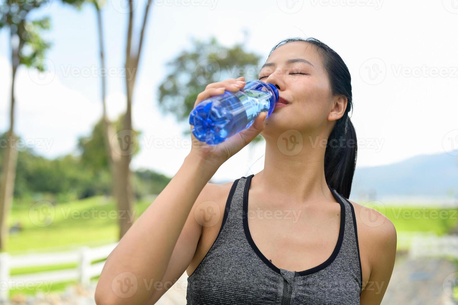 A fit woman in sportswear drinks from a water bottle, quenching her thirst after jogging in a park. photo