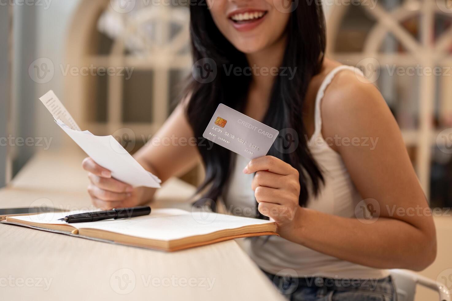 A close-up of a happy Asian woman holding a credit card and reviewing receipts at a table indoors. photo