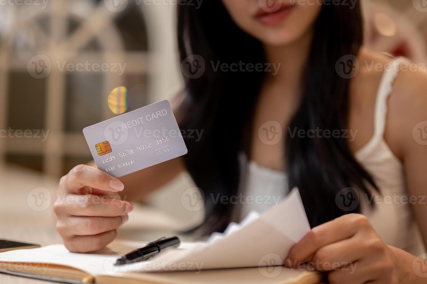 A thoughtful Asian woman holding a credit card and reviewing receipts at a table indoors. photo