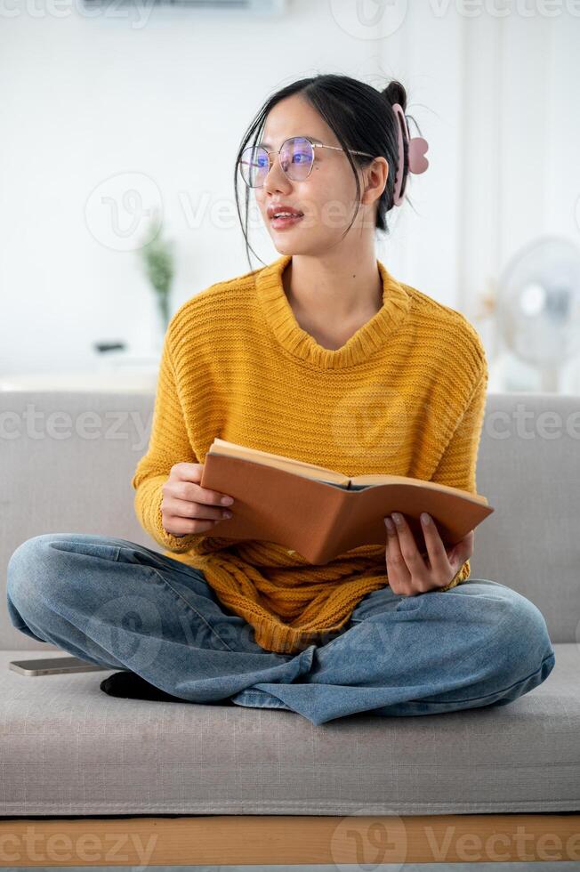 A beautiful Asian woman is daydreaming while looking out the window and enjoying reading a book. photo