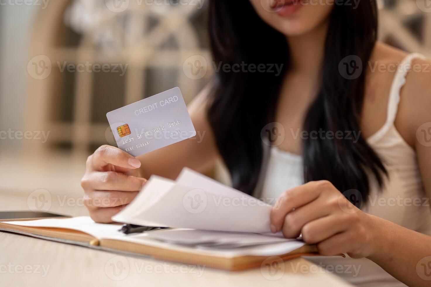 A thoughtful Asian woman holding a credit card and reviewing receipts at a table indoors. photo