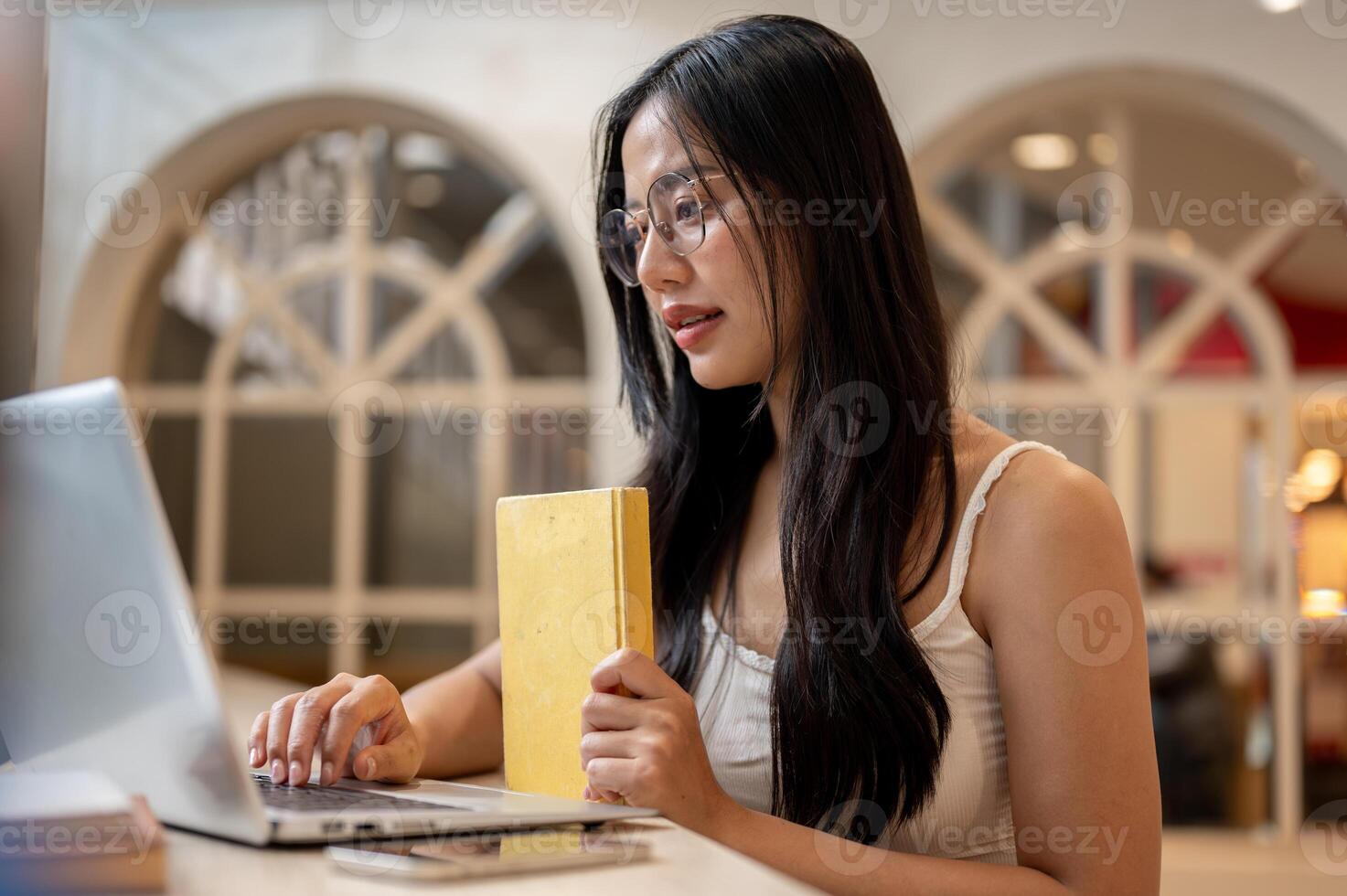 A woman is focused on working on her laptop, sitting in a library or a cafe. photo