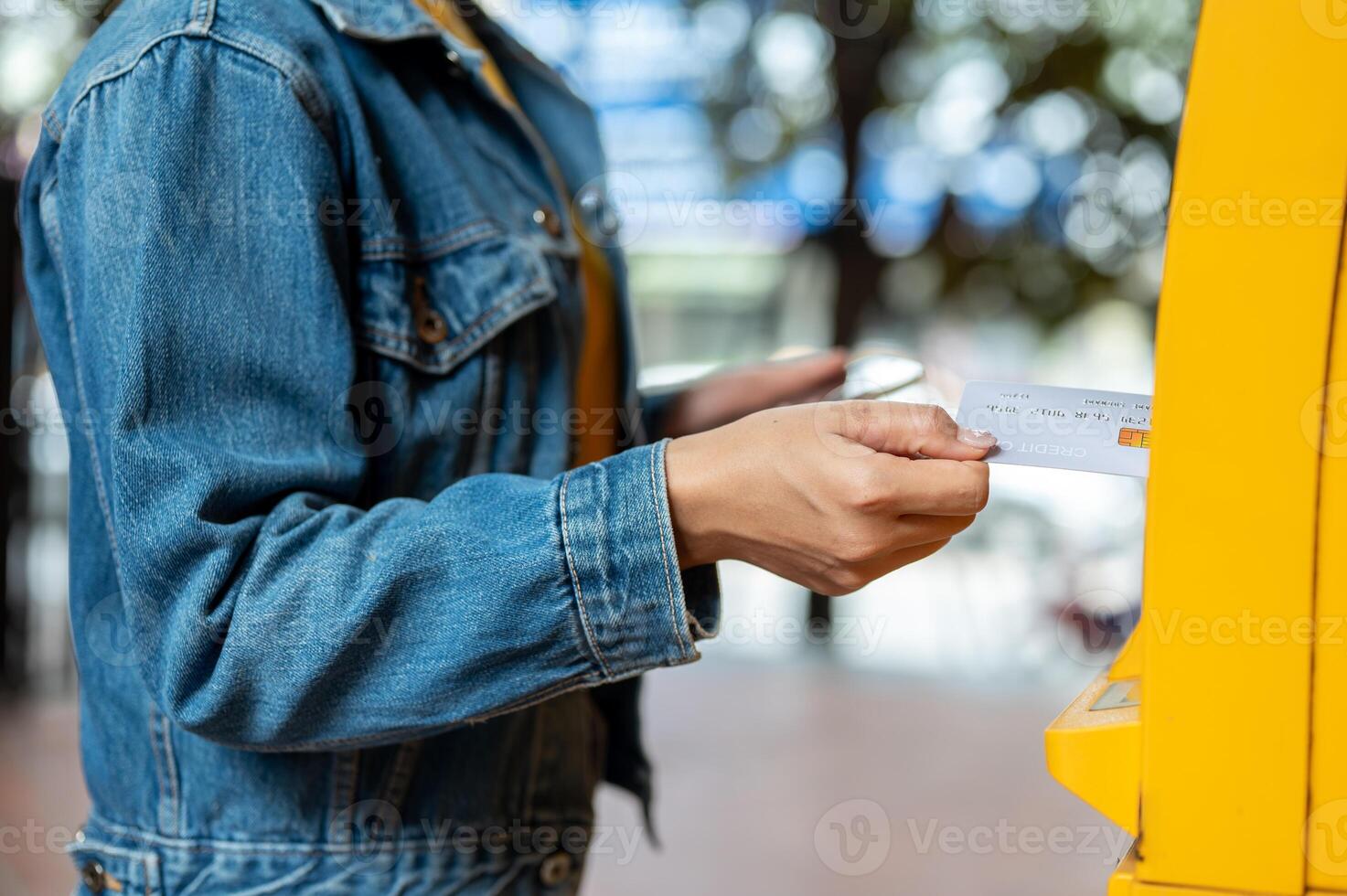A close-up of a woman in a denim jacket inserting her debit card into an ATM to withdraw some money. photo