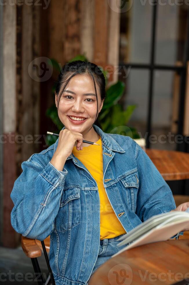 A charming, positive Asian woman in a denim jacket is reading a book while sitting in a coffee shop. photo