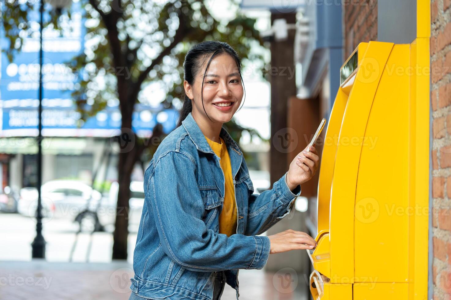 A beautiful Asian woman in a denim jacket is standing in front of an ATM, smiling at the camera. photo
