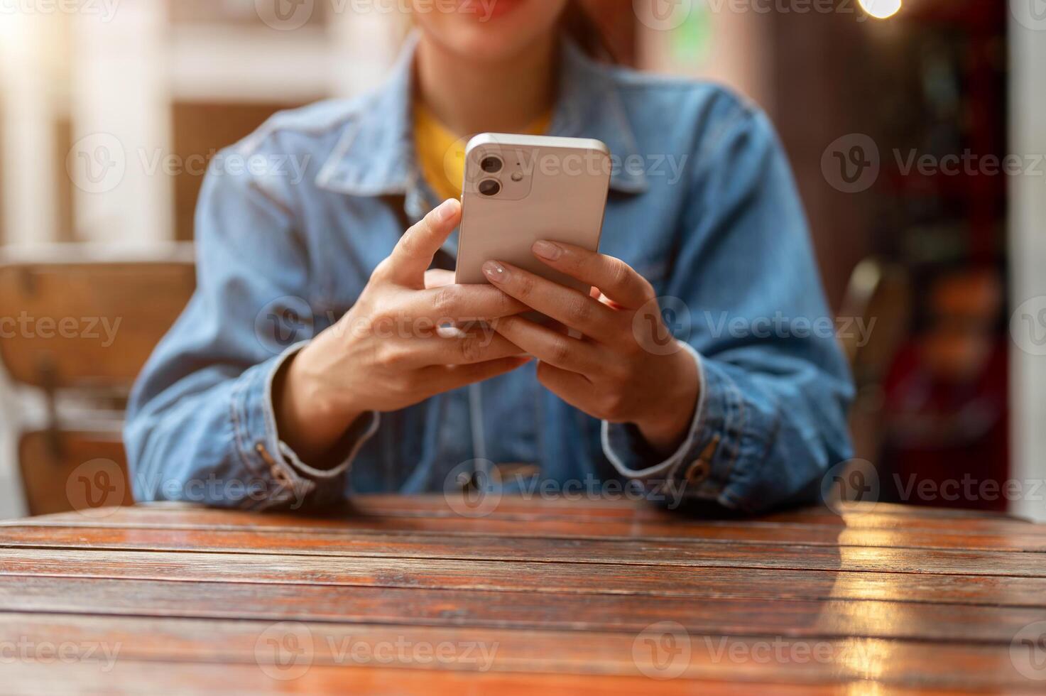 A cropped image of a woman in a denim jacket using smartphone while sitting at a table outdoors. photo