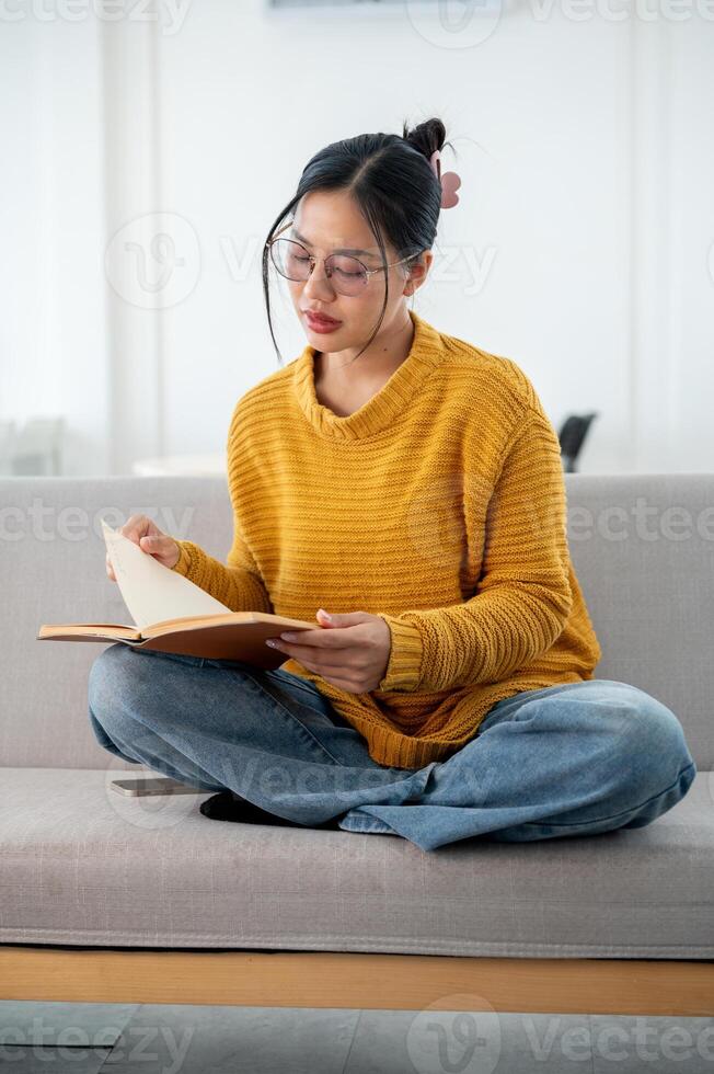 A beautiful Asian woman in casual clothes focuses on reading a book on a couch at home. photo